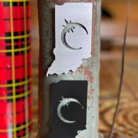 Close-up of a rusted metal Indiana Solar Eclipse Magnet with cut-out crescent moon and star designs, standing on a patterned tablecloth.